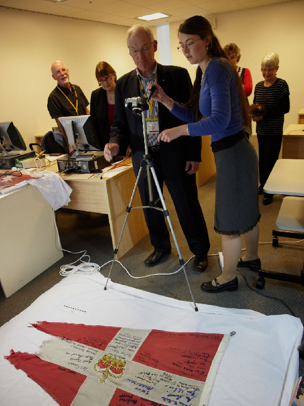 Museum volunteers standing around a camera on a tripod photographing a torn red and white flag with signatures on it